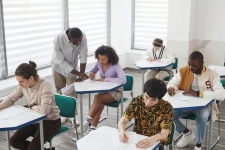 Advanced Placement Source resource image showing a classroom full of students with a teacher leaning over the desk of one student and helping her with her work