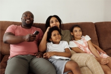 Hoopla resource image showing a young family sitting on the couch and watching tv, A father, mother, and two daughters